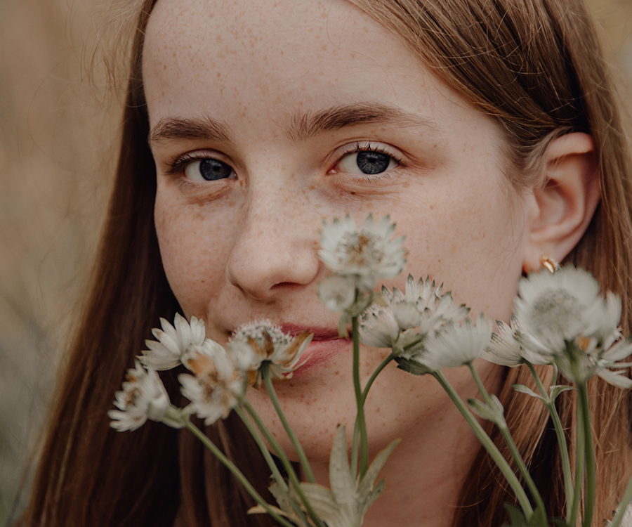 Junge Frau mit Sommersprossen schaut verträumt durch weiße Blumen in die Kamera